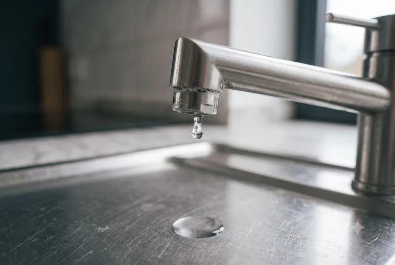 Einzelner Wassertropfen am Auslauf einer Küchenarmatur, darunter unscharf das Edelstahlbecken der Spüle
