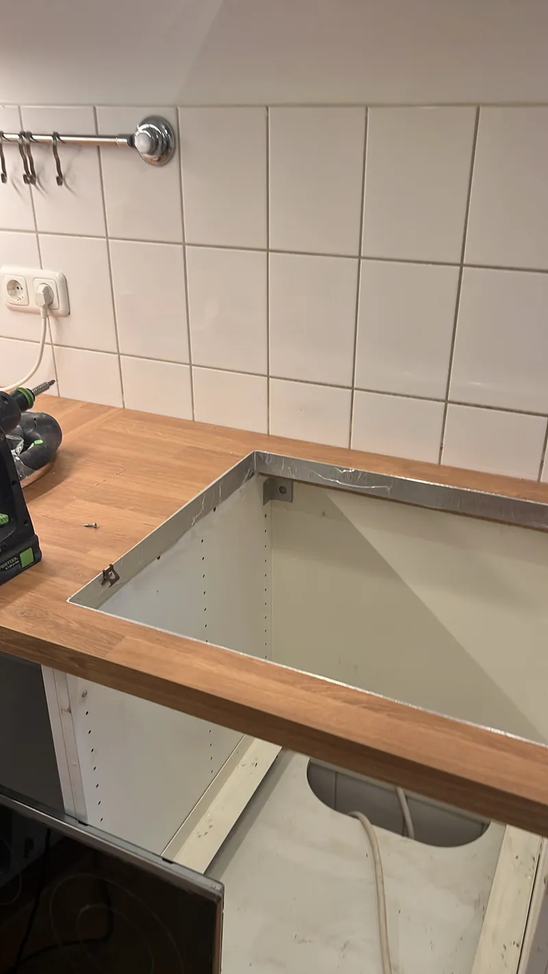 Rechteckiger Spülenausschnitt in einer neuen hellen Eiche-Arbeitsplatte, die Schnittkante ist umlaufend mit Dichtband versiegelt, darunter der offene Unterschrank, links ein Akkuschrauber.