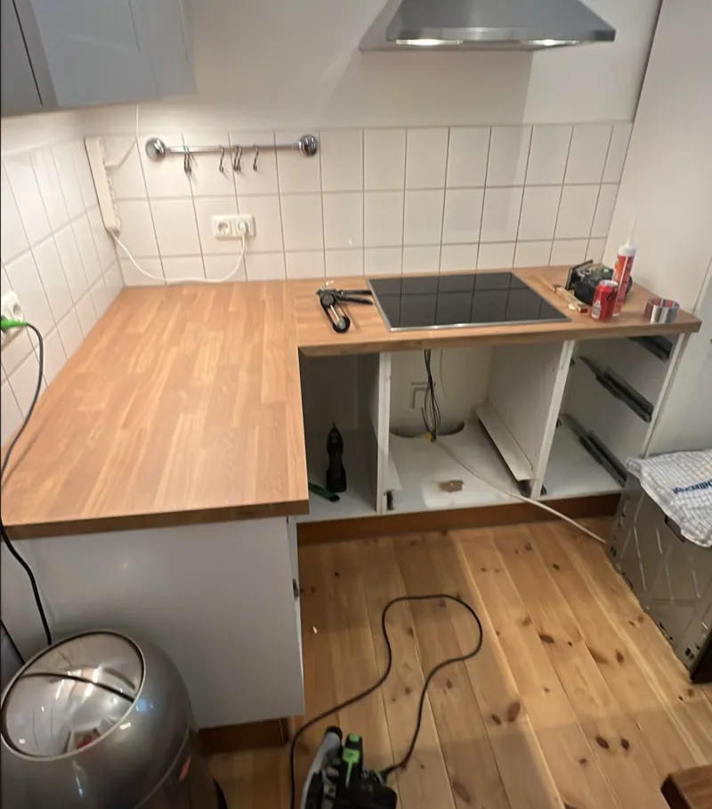 Neue helle Eiche-Arbeitsplatte liegt über der über Eck geführten Küchenzeile, das schwarze Kochfeld sitzt im Ausschnitt, die Unterschränke darunter sind noch offen.